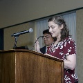 An individual stands at a podium with a microphone, raising one hand while reading from a program during the Alpha Omega Alpha 2023 Induction Ceremony at UTMB, with another attendee visible behind.