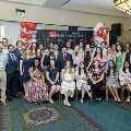 A large group of attendees pose for a formal group photo at the Alpha Omega Alpha 2023 Induction Ceremony at UTMB in a ballroom with patterned carpet, chandeliers, and a UTMB backdrop.