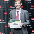 An individual holds a certificate while posing at the Alpha Omega Alpha 2023 Induction Ceremony at UTMB, standing in front of a UTMB Health and John Sealy School of Medicine logo backdrop.
