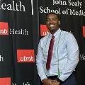 An individual poses at the Alpha Omega Alpha 2023 Induction Ceremony at UTMB, standing in front of a UTMB Health and John Sealy School of Medicine branded backdrop.