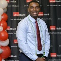 An individual poses at the Alpha Omega Alpha 2023 Induction Ceremony at UTMB, standing in front of a UTMB Health and John Sealy School of Medicine logo backdrop with a column of red, white, and gray balloons to the side.