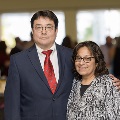 Two individuals stand together at the Alpha Omega Alpha 2023 Induction Ceremony at UTMB in a reception area with soft lighting, catered items, and other attendees visible in the background.