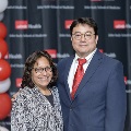 Two individuals pose together at the UTMB Alpha Omega Alpha Texas Chapter Fall Mixer, standing in front of a black logo backdrop. One wears a patterned top and glasses, while the other wears a blue suit with a red tie.