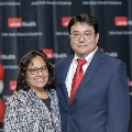 Two attendees pose together at the UTMB Alpha Omega Alpha Texas Chapter Fall Mixer in front of a black event backdrop with repeating UTMB Health logos. One wears a patterned top and glasses; the other wears a blue suit with a red tie.
