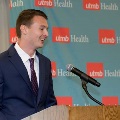 Individual in a blue suit stands at a wooden podium with a microphone in front of a UTMB Health backdrop during an Alpha Omega Alpha Texas Alpha Chapter event.
