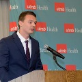 Individual in a blue suit speaks at a wooden podium with a microphone in front of a UTMB Health backdrop during an Alpha Omega Alpha Texas Alpha Chapter event.