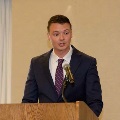 Individual in a blue suit stands at a wooden podium with a microphone, speaking during an Alpha Omega Alpha Texas Alpha Chapter event at UTMB.