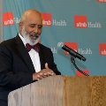 Individual in a suit with a bow tie stands at a wooden podium speaking into a microphone in front of a UTMB Health backdrop during an Alpha Omega Alpha Texas Alpha Chapter event.