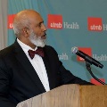 Individual in a dark suit with a bow tie stands at a wooden podium speaking into a microphone in front of a UTMB Health backdrop during an Alpha Omega Alpha Texas Alpha Chapter event.