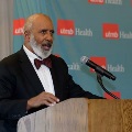 Individual in a dark suit with a bow tie speaks at a wooden podium with a microphone in front of a UTMB Health backdrop during an Alpha Omega Alpha Texas Alpha Chapter event.