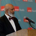 Individual in a dark suit with a bow tie speaks at a wooden podium with a microphone in front of a UTMB Health backdrop during an Alpha Omega Alpha Texas Alpha Chapter event.