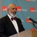 Individual in a dark suit with a bow tie speaks at a wooden podium with a microphone in front of a UTMB Health backdrop during an Alpha Omega Alpha Texas Alpha Chapter event.