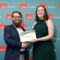 Two individuals stand in front of a UTMB Health backdrop while holding a certificate together during an Alpha Omega Alpha Texas Alpha Chapter event.