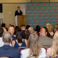 Speaker stands at a podium addressing a seated audience in rows of red chairs in front of a UTMB Health backdrop during an Alpha Omega Alpha Texas Alpha Chapter event.