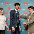 Individual places a green honor cord around another attendee wearing a suit in front of a UTMB Health backdrop, with a third attendee observing during an Alpha Omega Alpha Texas Alpha Chapter ceremony.