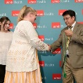 Two attendees shake hands while one holds green honor cords in front of a UTMB Health backdrop, with another attendee nearby during an Alpha Omega Alpha Texas Alpha Chapter ceremony.