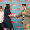 Two attendees shake hands while one holds green honor cords in front of a UTMB Health backdrop, with another attendee nearby during an Alpha Omega Alpha Texas Alpha Chapter ceremony.