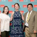 Three individuals pose indoors in front of a UTMB Health and John Sealy School of Medicine branded backdrop, with one wearing green honor cords, all dressed in semi-formal attire and standing close together for a photo.