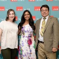 Three individuals pose indoors in front of a UTMB Health and John Sealy School of Medicine branded backdrop, with one wearing green honor cords, all dressed in semi-formal attire and standing close together.