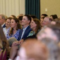 Audience members sit in rows during an indoor ceremony, facing forward attentively, with chairs arranged closely together and a softly lit event space visible in the background.