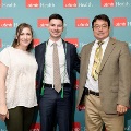 Three individuals pose indoors in front of a UTMB Health and John Sealy School of Medicine branded backdrop, with one wearing green honor cords, all dressed in semi-formal attire and standing close together for a photo.