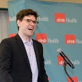 Individual in a dark suit stands at a podium speaking into a microphone in front of a UTMB Health branded backdrop, with a teal background and repeated “utmb Health” logos in a softly lit indoor event space.