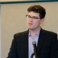 Individual in a suit stands behind a podium speaking into a microphone during an indoor ceremony, with a neutral background and presentation screen visible behind.