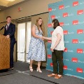 Individual in a patterned dress shakes hands with another individual near a small stage, with a UTMB Health branded backdrop, podium, and curtain visible in a softly lit indoor ceremony setting.