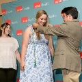 Individual in a patterned dress stands still as another individual places green honor cords over the shoulders in front of a UTMB Health branded backdrop, with a third person observing nearby.