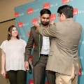 One individual places green honor cords over another individual’s shoulders in front of a UTMB Health branded backdrop, while a third person stands nearby in semi-formal attire during the indoor ceremony.