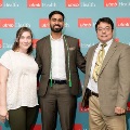 Three individuals pose indoors in front of a UTMB Health and John Sealy School of Medicine branded backdrop, with one wearing green honor cords, all dressed in semi-formal attire and standing close together for a photo.