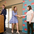 Individual in a blue patterned dress shakes hands with another individual near a small stage, with a UTMB Health branded backdrop and podium visible nearby in a softly lit ceremony setting.