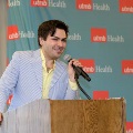 Individual stands at a wooden podium speaking into a microphone, with a UTMB Health branded backdrop behind and notes placed on the podium in a softly lit indoor ceremony setting.
