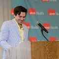 Individual stands at a wooden podium speaking into a microphone, with a UTMB Health branded backdrop behind and notes placed on the podium in a softly lit indoor ceremony setting.