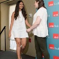 Individual in a white dress steps onto a small stage and extends a hand to greet another individual in front of a UTMB Health branded backdrop, with a curtain and platform edge visible in an indoor ceremony setting.