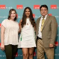 Three individuals pose indoors in front of a UTMB Health and John Sealy School of Medicine branded backdrop, with repeating logos, dressed in semi-formal attire and standing close together for a photo.