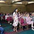 Individual in a white floral dress walks down an aisle between seated attendees during an indoor ceremony, with rows of chairs, chandeliers, and a patterned carpet visible in the event space.