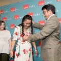 Individual in a floral dress stands in front of a UTMB Health branded backdrop as another individual places green honor cords over the shoulders, with a third person standing nearby during an indoor ceremony.