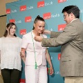 One individual places green honor cords over another individual’s shoulders in front of a UTMB Health branded backdrop, while a third person stands nearby in semi-formal attire during an indoor ceremony.