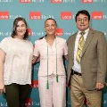 Three individuals pose indoors in front of a UTMB Health and John Sealy School of Medicine branded backdrop, one wearing green honor cords, all dressed in semi-formal attire and standing close together.
