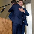 Two individuals in suits embrace on a small stage near a wooden podium with a microphone, during an indoor ceremony with neutral walls and soft lighting.