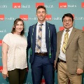 Three individuals pose indoors in front of a UTMB Health and John Sealy School of Medicine branded backdrop, with one wearing green honor cords, all dressed in semi-formal attire and standing close together.