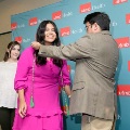 Individual wearing a bright pink outfit stands in front of a UTMB Health branded backdrop as another individual places green honor cords over the shoulders, with a third person visible nearby in a formal ceremony setting.