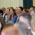 Audience members sit in rows during an indoor ceremony, facing forward attentively, with a softly lit event space, chairs, and neutral-colored walls visible in the background.