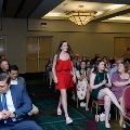 Individual in a red dress walks down an aisle between seated attendees during an indoor ceremony, with rows of chairs, a patterned carpet, and chandeliers visible overhead.