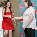 Individual in a red dress steps onto a small stage and reaches forward to shake hands with another individual near a UTMB Health branded backdrop, with a microphone stand and curtain visible.