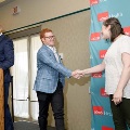 Two individuals shake hands near a stage during an indoor ceremony, with a UTMB Health branded backdrop and a podium visible nearby, while another presenter stands at the podium in a softly lit event space.