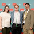 Three individuals pose indoors in front of a UTMB Health and John Sealy School of Medicine branded backdrop, with repeating red logo panels, dressed in semi-formal attire and standing close together for a group photo.