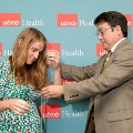 One individual places green honor cords over another individual’s shoulders in front of a UTMB Health and John Sealy School of Medicine branded backdrop, both standing indoors in semi-formal attire during a ceremony.