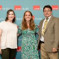 Three individuals pose indoors in front of a UTMB Health and John Sealy School of Medicine branded backdrop, with repeating logos, dressed in semi-formal attire and standing close together for a photo.